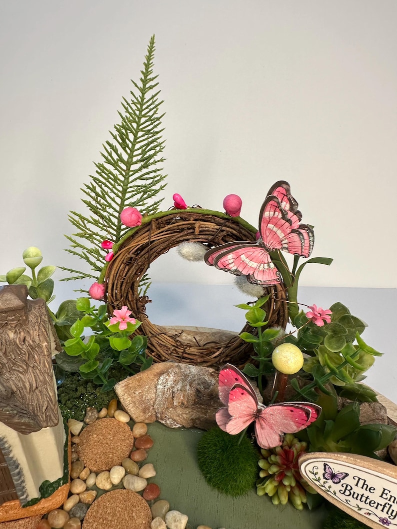 K&ouml;nnte beinhalten: Eine Miniatur-Feengarten-Szene mit einem braunen Zweigkranz mit rosa Akzenten, einem kleinen Haus und dekorativen Schmetterlingen. Die Szene umfasst k&uuml;nstliches Gr&uuml;n, kleine Steine und ein Schild mit der Aufschrift "The Enchanted Butterfly".