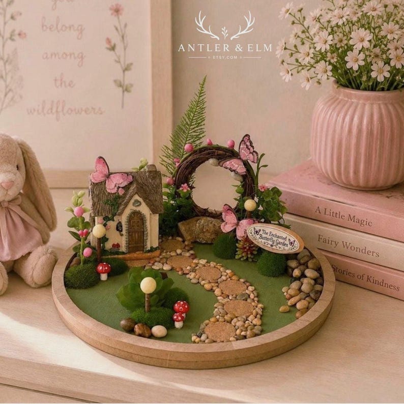 K&ouml;nnte beinhalten: Eine Miniatur-Feengarten-Szene mit einem kleinen Haus, Schmetterlingen und einem Steinweg. Die Szene befindet sich auf einer Holzbasis mit einer gr&uuml;nen Oberfl&auml;che, umgeben von kleinen Steinen. Eine rosa Vase mit wei&szlig;en Blumen und B&uuml;chern befindet sich im Hintergrund.