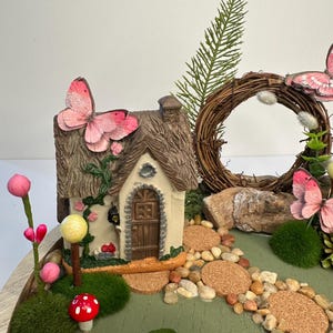 K&ouml;nnte beinhalten: Eine Miniatur-Feengarten-Szene mit einem kleinen H&auml;uschen mit Strohdach, einem Steinweg und dekorativen Elementen wie Schmetterlingen, Pilzen und einem Kranz. Die Farbpalette umfasst Gr&uuml;n-, Braun- und Rosat&ouml;ne.