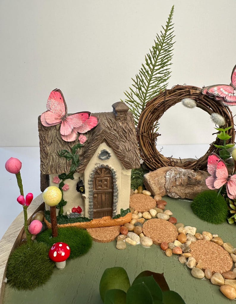 K&ouml;nnte beinhalten: Eine Miniatur-Feengarten-Szene mit einem kleinen H&auml;uschen mit Strohdach, einem Steinweg und dekorativen Elementen wie Schmetterlingen, Pilzen und einem Kranz. Die Farbpalette umfasst Gr&uuml;n-, Braun- und Rosat&ouml;ne.