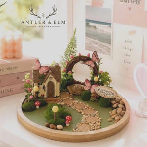 K&ouml;nnte beinhalten: Eine Miniatur-Feengarten-Szene mit einem kleinen Haus, einem Steinweg und einem Kranz mit Schmetterlingen. Die Szene befindet sich auf einem Holztablett mit gr&uuml;nem Untergrund und kleinen Steinen. Der Text "The Enchanted Butterfly Garden" ist sichtbar.