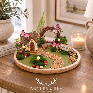 K&ouml;nnte beinhalten: Eine Miniatur-Feengarten-Szene mit einem kleinen Haus, einem Steinweg und einem Kranz mit rosa Schmetterlingen. Die Szene befindet sich auf einem Holztablett, mit einer gr&uuml;nen Basis und kleinen Pilzen.