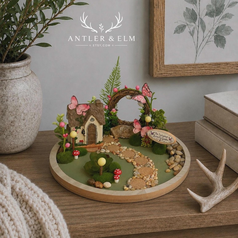 K&ouml;nnte beinhalten: Eine Miniatur-Feengarten-Szene mit einem kleinen H&auml;uschen, einem Steinweg und rosa Schmetterlingen. Die Szene befindet sich auf einer runden Holzbasis mit gr&uuml;ner Oberfl&auml;che, umgeben von einem Holzrahmen. Der Text "The Enchanted Butterfly Garden" ist sichtbar.