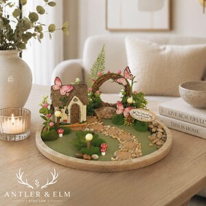 K&ouml;nnte beinhalten: Eine Miniatur-Feengarten-Szene mit einem reetgedeckten H&auml;uschen, einem Steinweg und dekorativen Schmetterlingen. Der Garten befindet sich auf einem Holztablett mit gr&uuml;ner Basis, umgeben von kleinen Steinen und Miniaturpflanzen. Die Szene enth&auml;lt ein kleines Schild mit der Aufschrift "The Enchanted Butterfly Garden".