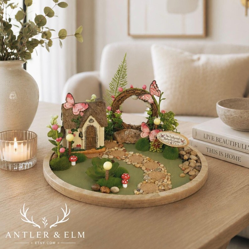 K&ouml;nnte beinhalten: Eine Miniatur-Feengarten-Szene mit einem reetgedeckten H&auml;uschen, einem Steinweg und dekorativen Schmetterlingen. Der Garten befindet sich auf einem Holztablett mit gr&uuml;ner Basis, umgeben von kleinen Steinen und Miniaturpflanzen. Die Szene enth&auml;lt ein kleines Schild mit der Aufschrift "The Enchanted Butterfly Garden".