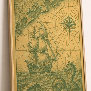 Op de afbeelding: Een ingelijste kunstdruk met een groene inktillustratie op een gouden achtergrond. Het kunstwerk toont een zeilschip op woelige golven, een zeemonster dat uit het water oprijst en een kompasroos met kruisende navigatielijnen.