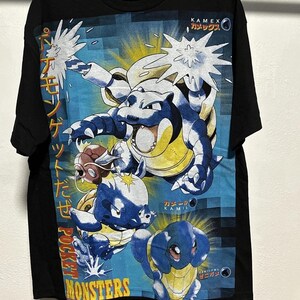 Puede incluir: Camiseta negra con un diseño de monstruos de dibujos animados azules y amarillos con texto japonés. El diseño incluye las palabras "POCKET MONSTERS" y "KAMEX". La camiseta tiene cuello redondo y mangas cortas.