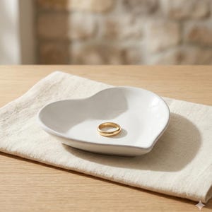 Pode incluir: Uma tigela de anel de cerâmica branca em forma de coração com um anel dourado dentro. A tigela está sobre uma toalha de linho bege, colocada sobre uma superfície de madeira clara. O fundo apresenta uma parede de tijolos.