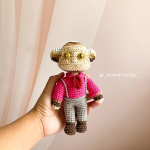 Puede incluir: Un muñeco de mono tejido a ganchillo con cabeza beige y detalles marrones, ojos amarillos, camisa rosa brillante con corbata roja, pantalones grises con tirantes, y manos y pies marrones. El texto @_mimirricha_ es visible a la derecha.