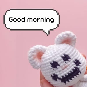 Puede incluir: Un juguete de peluche tejido a ganchillo en color blanco con orejas redondas y una cara negra cosida que muestra una amplia sonrisa. Sobre el juguete, una burbuja de diálogo blanca con borde negro pixelado contiene el texto "Good morning".