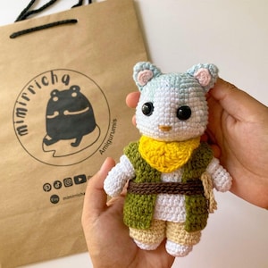 Könnte beinhalten: Eine gehäkelte Tierfigur mit hellblau-weißem Kopf, schwarzen Augen und rosa Innenohren. Sie trägt einen gelben Halsschal, eine grüne Weste, einen braunen Gürtel und eine beigefarbene Hose. Die Figur ist vor einer braunen Papiertüte mit dem Text mimirricha.