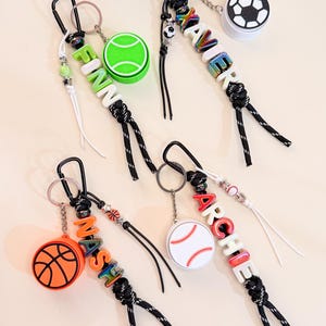 Könnte beinhalten: Vier sportliche Schlüsselanhänger mit schwarzen Karabinern und Kordeln. Jeder enthält einen Sportball-Anhänger – Tennis, Fußball, Basketball oder Baseball – sowie bunte Buchstabenperlen, die die Namen FINN, XAVIER, NASH und ARCHIE bilden.