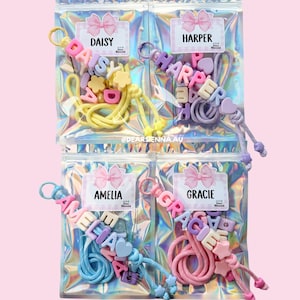 May include: Four holographic pouches containing DIY name keyrings with colourful letter beads, star and heart charms, and cords. Each pouch features a label with a pink bow graphic and the names Daisy, Harper, Amelia, or Gracie. Text reads: MAKE YOUR OWN NAME KEYRING and @DEARSIENNA.AU.