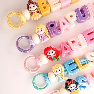 May include: Five colourful keychains featuring cartoon character charms and letter beads spelling out names like Belle, Rapunzel, Ariel, Elsa, and Snow White. Each keychain includes a metal lobster clasp, a round bead, and a cord with plastic letters.