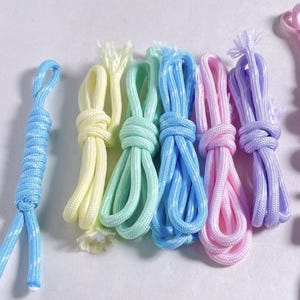 May include: A collection of braided cords in pastel colors including light blue, pale yellow, mint green, bright blue, soft pink, and lavender. Each cord is folded into a loop and features a woven texture with visible frayed ends at the tips.