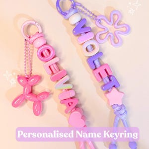 May include: Two personalised name keyrings featuring colourful plastic letter beads spelling OLIVIA and VIOLET. The OLIVIA keyring includes a pink balloon dog charm, while the VIOLET keyring features a purple flower charm and a star bead.