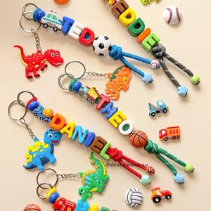 May include: Four personalised keychains featuring colourful letter beads spelling the names EDDIE, THEO, DANIEL, and HARRY. Each keychain includes various silicone charms shaped like dinosaurs, sports balls, and vehicles attached to metal rings.