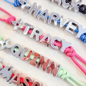 Può includere: Una collezione di portachiavi personalizzati con lettere in metallo argentato che compongono i nomi JACK, HARVEY, TRACY, QUINN e MASON. Ogni portachiavi è fissato a un cordino di nylon colorato e include un ciondolo in metallo a forma di stella.