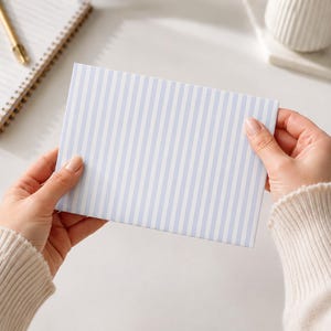 May include: A rectangular card or envelope featuring a vertical pinstripe pattern in light blue and white. The item is held horizontally, displaying a clean, uniform design with alternating thin blue stripes against a crisp white background.