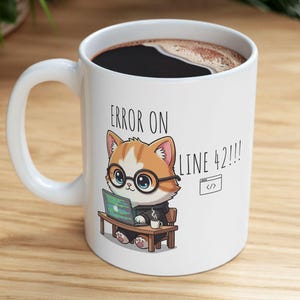 May include: White ceramic coffee mug with a handle, featuring a cartoon cat wearing glasses and working on a laptop. The mug has the text "ERROR ON LINE 42!!!" and a small code icon. The mug is filled with dark coffee.