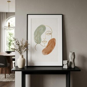 May include: Abstract art print featuring a minimalist line drawing of a face in gold against a white background. The artwork includes green and rust-colored watercolor shapes. The print is framed in a black frame.