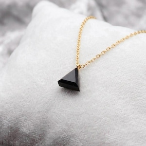 May include: A necklace featuring a small, black, triangular-shaped pendant suspended from a delicate gold-toned metal chain. The pendant has a smooth, faceted surface that reflects light, and the chain consists of small, uniform circular links.