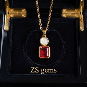 May include: A gold-tone necklace featuring a pendant with a rectangular red gemstone and a circular white mother-of-pearl accent. The pendant hangs from a delicate gold-tone chain. The text ZS gems is printed in gold below the pendant.