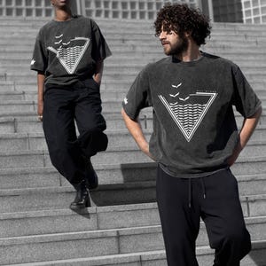 May include: Two people wearing matching dark grey t-shirts and black trousers are on a staircase. The t-shirts feature a white triangular design with waves and birds. The background is a grayscale urban setting.