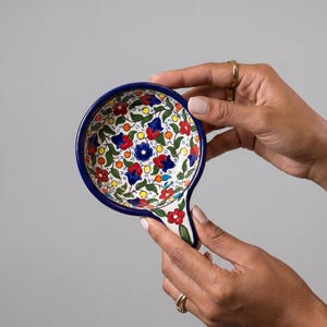 Puede incluir: Un pequeño reposacucharas redondo de cerámica con mango, que presenta un vibrante patrón floral pintado a mano en rojo, azul, amarillo y verde sobre un fondo blanco, con un borde azul oscuro sólido y una superficie esmaltada brillante.