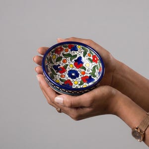 Könnte beinhalten: Eine kleine Keramikschale mit handgemaltem Blumenmuster in leuchtendem Rot, Blau und Gelb auf weißem Grund. Die Schale hat einen tiefblauen Rand und eine dekorative Borte aus dunkelblauen und türkisfarbenen geometrischen Formen am Boden.