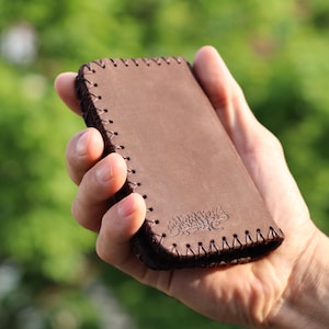 May include: A rectangular brown leather case featuring dark brown whipstitching along the edges. The surface of the leather displays a debossed Arabic calligraphy design in the lower corner, providing a textured detail to the smooth material.
