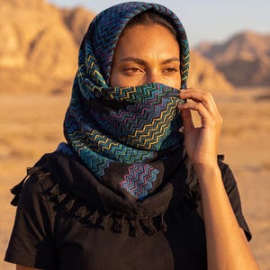 Puede incluir: Un pañuelo negro con un vibrante patrón de chevrón multicolor en tonos azules, verdes y morados. La tela está drapeada sobre la cabeza y alrededor del rostro, con borlas negras que bordean los extremos de este accesorio textil cuadrado.
