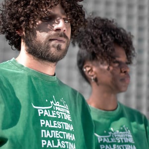 May include: Two individuals are seen wearing green sweatshirts. The sweatshirts feature the word "Palestine" in multiple languages, including Arabic, English, Russian, Spanish, Japanese, and Hindi, in white lettering. Both have dark curly hair.