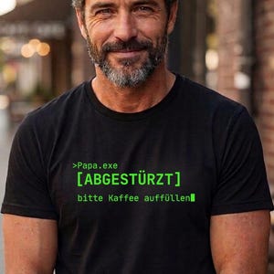 Op de afbeelding: Zwart T-shirt met groene tekst: '>Papa.exe [ABGESTÜRZT] bitte Kaffee auffüllen'. De tekst is in een lettertype in computercode-stijl. Het T-shirt wordt gedragen door een persoon met een baard.