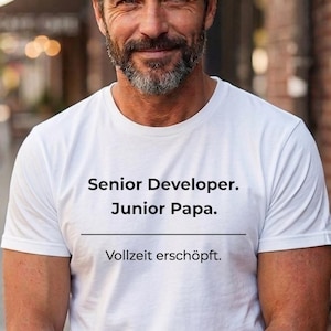 Könnte beinhalten: Weißes T-Shirt mit dem Text "Senior Developer. Junior Papa. Vollzeit erschöpft." in Schwarz gedruckt. Das T-Shirt wird von einer Person mit grauem Bart getragen.