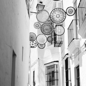 May include: Black and white photograph of a narrow street with white buildings and decorative circular lace ornaments suspended overhead. Balconies with black railings and barred windows add architectural detail.