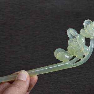 May include: A pale green, translucent hair stick featuring a carved floral design at one end. The ornament displays smooth, rounded petals and leaves sculpted from a single piece of material, with a long, tapered shaft extending from the base.