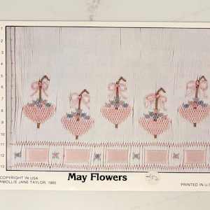 May include: A vintage needlepoint pattern titled "May Flowers." The design features five pink heart-shaped flowers with bows and blue accents, arranged above a row of rectangular shapes. The pattern is printed on a white background.