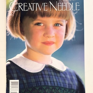 May include: A magazine cover featuring a young girl with short blonde hair and blue eyes. The magazine title "CREATIVE NEEDLE" is displayed in large, elegant font. The girl is wearing a blue and green plaid dress with a white collar. The cover also includes the date "July/Aug 1998".