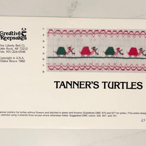 May include: A white card with the text "Creative Keepsakes" and "Tanner's Turtles." The card features a cross-stitch design of turtles in green and red. The design is stitched on a white background.
