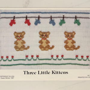 May include: A cross-stitch pattern featuring three brown kittens with red collars. Above them are red, blue, and green mittens hanging on a clothesline. The text "Three Little Kittens" is below the kittens.