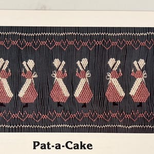 May include: A textile design featuring six figures in pink and white dresses with bonnets, set against a dark blue background. The design includes a decorative border and the text "Pat-a-Cake" at the bottom.
