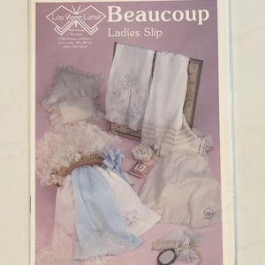 May include: A package featuring a variety of white and pastel-colored embroidered lingerie items. The package includes a slip, pillows, and other delicate items. The text "Beaucoup Ladies Slip" is visible on the package.