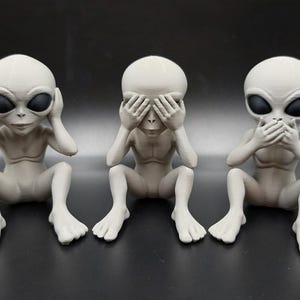 May include: Three light gray alien figurines in a seated position. The first alien covers its ears, the second covers its eyes with its hands, and the third covers its mouth. Each figure has a smooth, matte finish, large dark oval eyes, and a slender build.