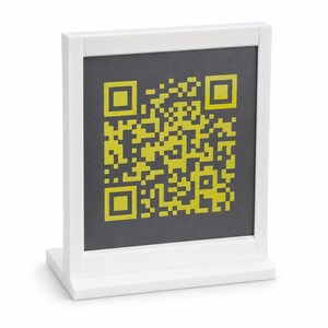 May include: A white rectangular sign holder featuring a dark gray square insert. The insert displays a bright yellow QR code pattern centered on the surface. The sign holder has a sturdy base and a clean, simple frame design for upright display.