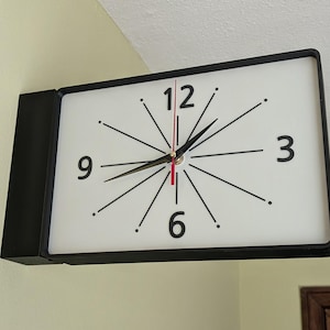 May include: A rectangular wall clock with a black frame and a white face. The clock features black numerals at the 3, 6, 9, and 12 positions, with black radiating lines and dots for the other hour markers. It has black hour and minute hands and a red second hand.