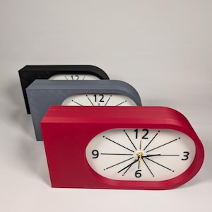 May include: Three horizontal table clocks with a rounded right side, featuring a white face with black hour markers, black hands, and the numbers 3, 6, 9, and 12. The clocks are arranged in a row, colored in red, gray, and black respectively.