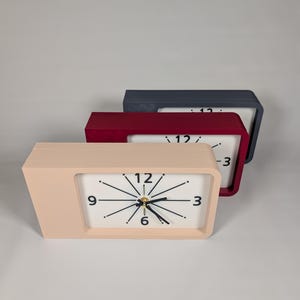 May include: Three rectangular desk clocks arranged in a staggered row. Each clock features a white face with black hands and black numerals at the 3, 6, 9, and 12 positions. The outer frames are solid colors: light pink, deep red, and dark gray.
