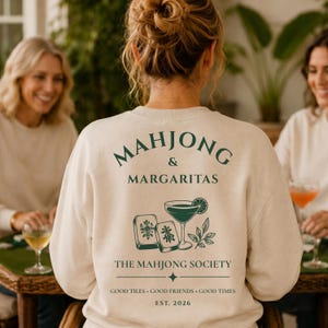 Peut inclure: Un sweat-shirt à col rond beige avec du texte et des graphismes vert foncé au dos. Le motif comprend les mots MAHJONG & MARGARITAS, THE MAHJONG SOCIETY, GOOD TILES GOOD FRIENDS GOOD TIMES, EST. 2026, ainsi que des illustrations de tuiles de mah-jong, un verre à cocktail et des feuilles.