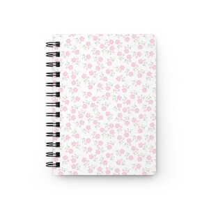 May include: A spiral-bound notebook with a white cover adorned with a delicate floral pattern. The design features small pink flowers and green leaves scattered across the surface. The notebook's binding is black.